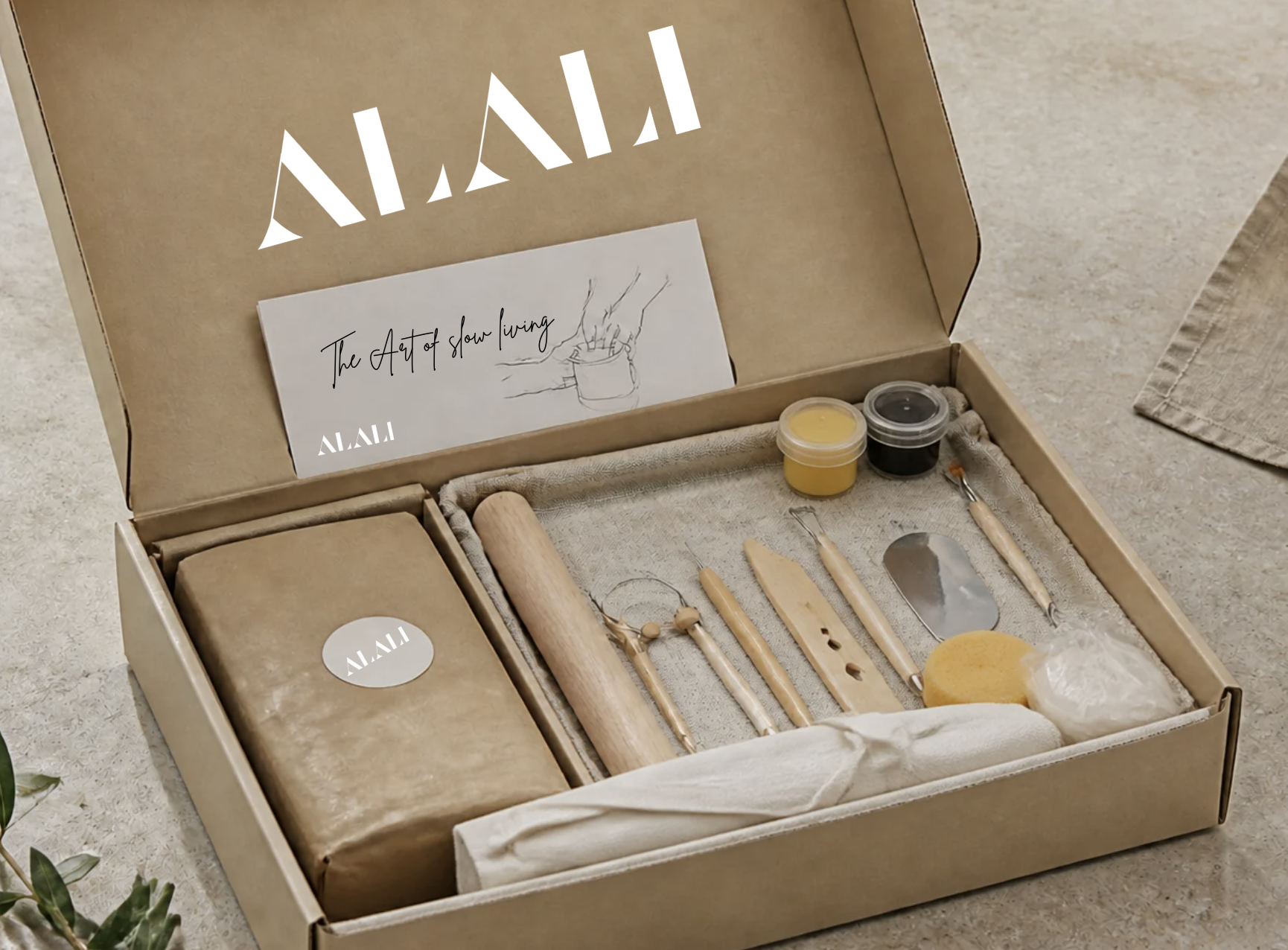 The Alali Clay Box opened on a table, with clay, tools, paints, gloves and an apron inside